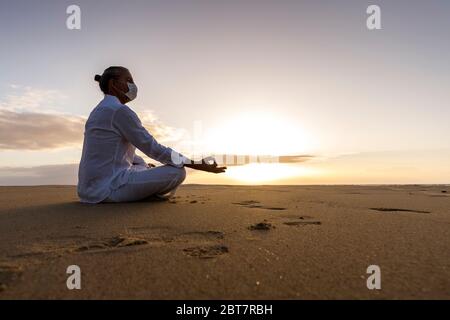 Meditierender Mann in medizinischer Maske in Lotushaltung am Strand, Männchen trägt chirurgische Gesichtsmaske und weiße Yoga-Kleidung mit Top-Knoten-Frisur bei Sonnenaufgang Stockfoto