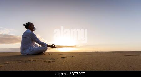 Meditierender Mann in medizinischer Maske in Lotushaltung am Strand, Männchen trägt chirurgische Gesichtsmaske und weiße Yoga-Kleidung mit Top-Knoten-Frisur bei Sonnenaufgang Stockfoto