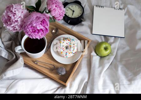Drei rote Pfingstrosen in einem weißen Porzellankrug, große Tasse Tee, Donut auf Untertasse, Teesieb, Apfel, auf Tablett, Wecker, Notizbuch auf Federn und ein Schlafzimmer Stockfoto