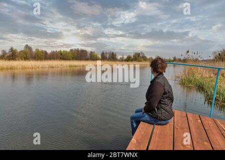 Kaukasische Frau mittleren Alters mit Blick auf den Fluss Ros, während sie auf einem Holzpflaster sitzt und ihre Beine umarmt, Ukraine. Stockfoto