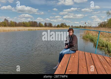 Kaukasische Frau mittleren Alters mit Blick auf den Fluss Ros, während sie auf einem Holzpflaster sitzt und ihre Beine umarmt, Ukraine. Stockfoto