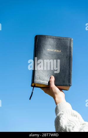 Erhobene Hand, die die Heilige Bibel hält. Hintergrund mit blauem Himmel an einem schönen Wintermorgen. Nahaufnahme. Kopierbereich. Vertikale Aufnahme. Stockfoto