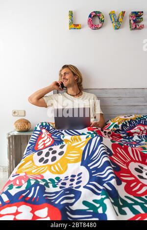 Junger Mann mit mittellangen blonden Haaren, mit Bart und Schnurrbart, der während der Quarantäne vom Bett aus von zu Hause aus am Computer arbeitet. Mit einem Kollegen sprechen Stockfoto