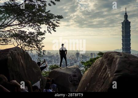 Taipei / Taiwan - 12. Dezember 2018: Silhouette eines jungen Mannes, der auf Felsen posiert, mit Blick auf den Wolkenkratzer Taipei 101 und das Stadtbild Taipehs vom Elephant Mountain Stockfoto