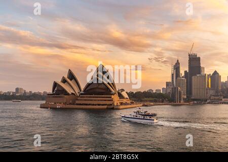 Sydney Opera House bei Sonnenuntergang. Wunderschöne orange-gelbe Farben. Die Stadt hinunter und der Himmel im Hintergrund trüb. Touristenboot im Vordergrund segeln. Stockfoto