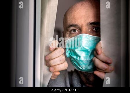 Der Mann, der medizinische Maske trägt, schaut durch das Fenster. Isolation zu Hause für Virenausbruch. Konzept Haus selbst Quarantäne, Prävention COVID-19. Stockfoto