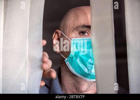 Der Mann, der medizinische Maske trägt, schaut durch das Fenster. Isolation zu Hause für Virenausbruch. Konzept Haus selbst Quarantäne, Prävention COVID-19. Stockfoto