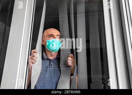 Der Mann, der medizinische Maske trägt, schaut durch das Fenster. Isolation zu Hause für Virenausbruch. Konzept Haus selbst Quarantäne, Prävention COVID-19. Stockfoto