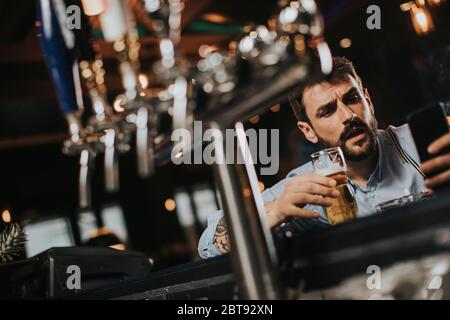 Junger Mann, der Bier trinkt, Zigarette raucht und im Pub Handy benutzt Stockfoto