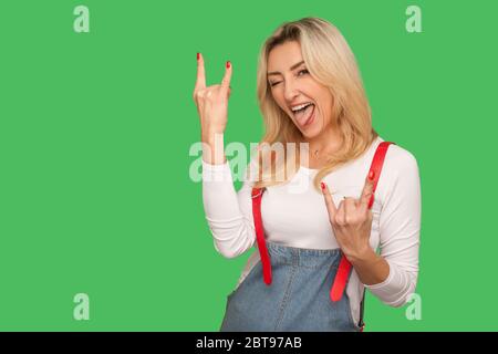 Das ist verrückt! Porträt der entzückenden erwachsenen Frau in stilvollen Denim Overalls zeigt Rock and Roll Geste und macht ungehorsame Grimasse mit der Zunge heraus Stockfoto