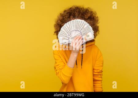 Lockige Frau im urbanen Stil Hoodie versteckt Gesicht hinter Haufen von Dollar-Banknoten, anonyme Person Geld halten, Lotterie gewinnen, großen Gewinn. Indoor Stockfoto