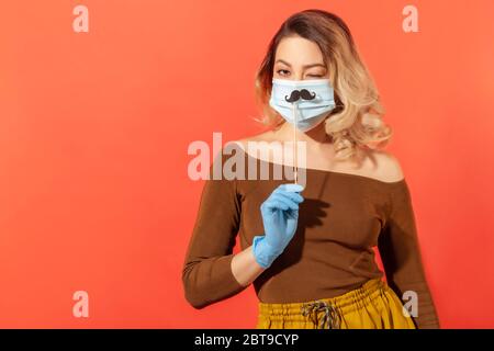 Porträt der positiven Frau in Schutzhandschuhen, hält gefälschte Schnurrbart Zubehör über chirurgische Gesichtsmaske, zwinkert spielerisch, in guter Laune trotz cor Stockfoto