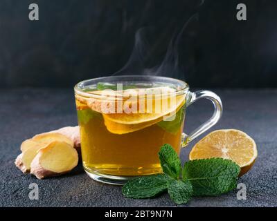Kräutertee mit Ingwer, Minze und Zitrone. Heißer Tee Glas Tasse mit Dampf und Zutaten auf schwarzem Zement Hintergrund. Kopierbereich Stockfoto