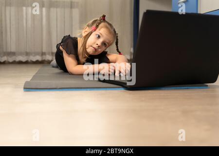 Ein kleines Mädchen in einem schwarzen Turntrikot macht Gymnastik zu Hause online vor einem Laptop. Platz für Text und Kopierer. Stockfoto