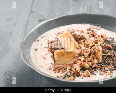 Nahaufnahme der Übernachtungen Hafer in der Schüssel mit Banane, LSA, Chia Samen, Mandeln und Honig auf grau Holztisch Hintergrund. Gesundes Frühstück Haferflocken Rezept Idee Stockfoto