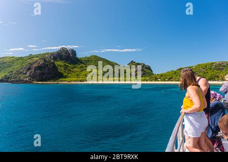 Touristen, die am Geländer einer Fähre stehen, blicken in der Ferne über das tiefblaue Meer auf eine tropische Insel in Fidschi Stockfoto