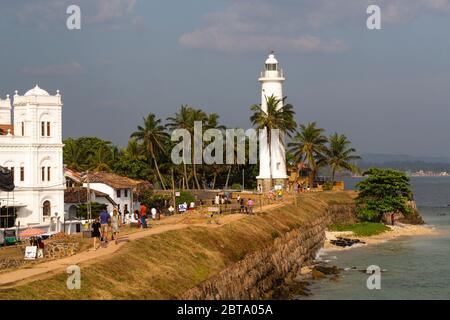 Die Touristen spazieren entlang der Stadtmauern von Galle Fort inmitten archäologischer Ausgrabungen. Ein von Palmen umgebenes Leuchtturm schützt die Hafenmündung. Stockfoto