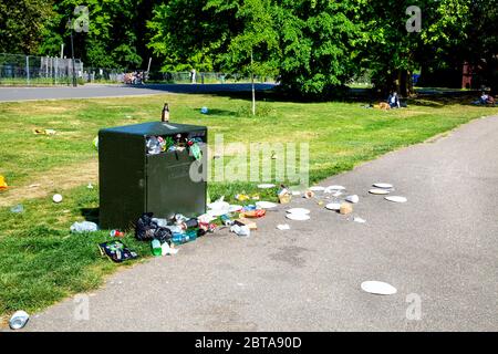 Ein Mülltonne in einem Park, der überfüllt und voller Müll ist, Müll auf dem Rasen und auf dem Weg verstreut (Battersea Park, London, UK) Stockfoto