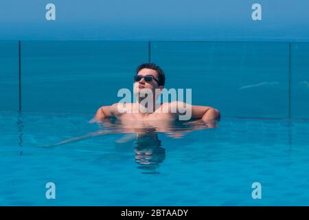 Junger kaukasischer Mann trägt Sonnenbrille am Rand eines Infinity-Pools mit blauem Meer und Himmel im Hintergrund. Lizenzfreies Stock Foto. Stockfoto