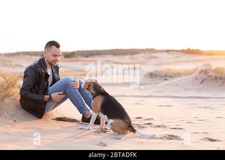 Junger kaukasischer Mann mit schwarzer Lederjacke und blauer Jeans sitzt am Strand neben seinem Freund, dem Hund Beagle Breed. Gutaussehender europäischer Typ Wal Stockfoto