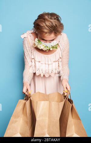 Mode Schönheit cool junge Mädchen in medizinische Maske mit entworfen Natürliche Blumen stehen mit Einkaufstaschen im Sommer Kleid über Blaue Wand Stockfoto