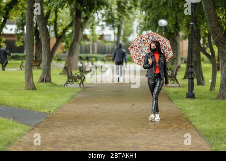 Ein maskiertes Mädchen läuft auf der Straße. Ein Mädchen in einer Schutzmaske geht im Park mit einem Regenschirm im Regen. Coronavirus-Infektion COVID-19 Stockfoto