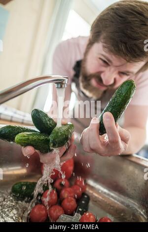Ein sorgfältiger Mann untersucht und wäscht Gurken unter einem Wasserstrom, Tomaten, Paprika und einem Messer in der Spüle in der Küche. Stockfoto