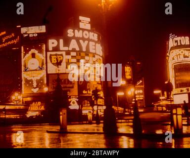 Die Neonschilder des Piccadilly Circus spiegeln sich im nächtlichen Regen auf der Straße wider und werben für die beliebten Produkte und Filme der 1960er Jahre, aufgenommen 1961. Stockfoto