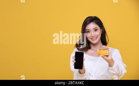Portrait Asian schöne glückliche junge Frau lächelnd fröhlich und sie ist mit Kreditkarte und mit Smartphone für Online-Shopping auf gelben Backgro Stockfoto