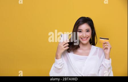 Portrait Asian schöne glückliche junge Frau lächelnd fröhlich und sie ist mit Kreditkarte und mit Smartphone für Online-Shopping auf gelben Backgro Stockfoto