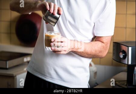 Mann gießt aufgeschäumte Milch in Espresso, um einen Latte Cappuccino Kaffee zuzubereiten. Lifestyle-Konzept für den Barista im Haus Stockfoto