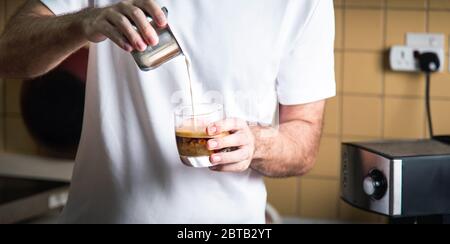 Mann gießt aufgeschäumte Milch in Espresso, um einen Latte Cappuccino Kaffee zuzubereiten. Lifestyle-Konzept für den Barista im Haus Stockfoto