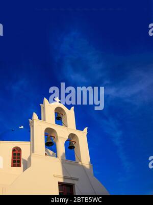 Santorini, Griechenland. Weiße orthodoxe Kirche und Glocken vor blauem Hintergrund am klaren Himmel, sonniger Frühlingsnachmittag, vertikales Foto, niedrige Winkelansicht Stockfoto