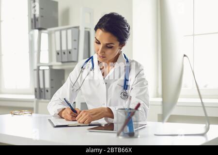 Selbstbewusste Frau Ärztin Kinderärztin schreibt in einer Zwischenablage, die an einem Tisch in einem weißen Büro des Krankenhauses sitzt. Stockfoto