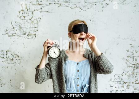 Schläfrig Mädchen in einer Schlafmaske hält einen Wecker in der Hand und gähnt. Stockfoto