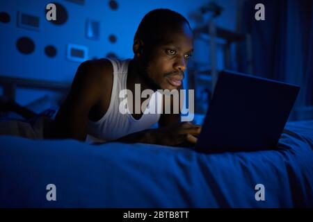 Porträt des jungen afroamerikanischen Mannes mit Laptop während auf dem Bett in der Nacht, kopieren Raum Stockfoto