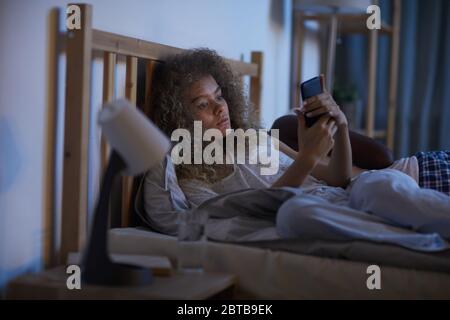 Porträt der jungen Lockenhaarigen Frau mit Smartphone im Bett in der Nacht, während sie an Schlaflosigkeit leiden, kopieren Raum Stockfoto
