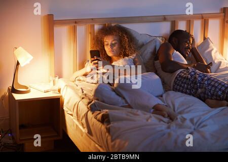 Porträt des jungen Mischrassenpaares mit Smartphones im Bett, während Rücken an Rücken getrennt liegen, kopieren Raum Stockfoto