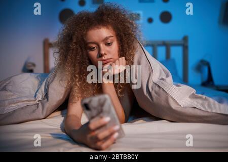 Vorderansicht Porträt von gelangweilten jungen Frau mit Smartphone im Bett in der Nacht von Decke bedeckt, kopieren Raum Stockfoto