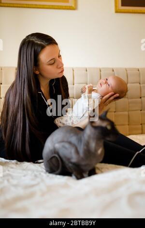 Sphynx Katze sitzt nahe Mutter und Baby Kleinkind auf weißem Bett Stockfoto