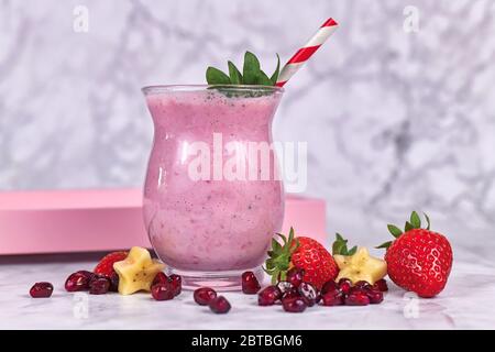 Smoothie-Shake aus rosa Früchten in Trinkglas mit gestreiftem Stroh, umgeben von Zutaten wie Erdbeer- und Bananenstücken und Granatapfelkernen Stockfoto