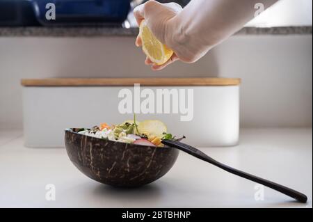 Weibliche Hand quetschen einen Zitronensaft auf köstlichen veganen gemischten Salat in natürlichen Kokosnussschüssel serviert. Stockfoto