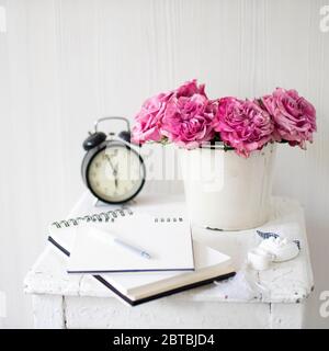Ein Strauß rosa Rosen in einem Zinn weißen Eimer auf einem alten Hocker mit Wecker und einer Serviette mit Stickerei neben dem Bett auf einem beigen Hintergrund Stockfoto