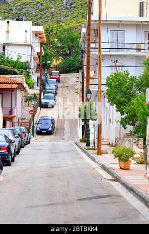 Eine schmale, typische alte Straße von Loutraki in Griechenland mit einer gesprungenen Straße inmitten geparkter Autos und grüner Bäume am frühen Morgen. Stockfoto