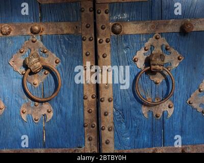 Detail von primitiven Gesicht Designs auf Metall Tür Klopfer auf blau lackiert traditionelle Doppeltür mit besetzt Metallarbeiten in Malindi, Kenia Küste, Afrika Stockfoto