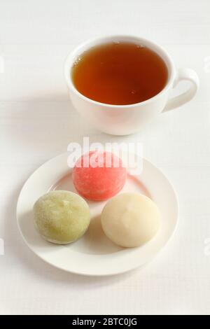 Drei Arten japanischer Dessertmochi - Granatapfel mit Honig, grüner Matcha-Tee, Kokosnuss auf einer weißen Untertasse und eine Tasse mit Tee auf einem weißen Holztisch Stockfoto