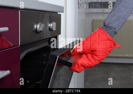 Eine Frauenhand mit roter Hitze schützt die Küchenhandschuhe beim Öffnen des Backofens. Essen und Speisen im Küchenkonzept zubereiten. Nahaufnahme, selektiver Fokus Stockfoto