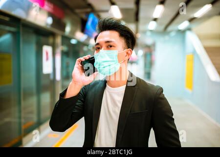 Junger Geschäftsmann mit Gesichtsmaske, der am Telefon telefoniert und auf den Zug wartet Stockfoto