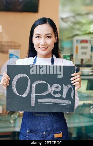Positive junge asiatische Frau in denin Schürze im Café stehen und zeigt offene Zeichen Stockfoto
