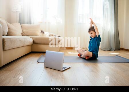 Der Junge macht körperliche Übungen, sieht Video-Lektion online vor offenem Laptop auf dem Boden. Körperliche Übungen auf Distanz. Quarantäne zu Hause. Stockfoto